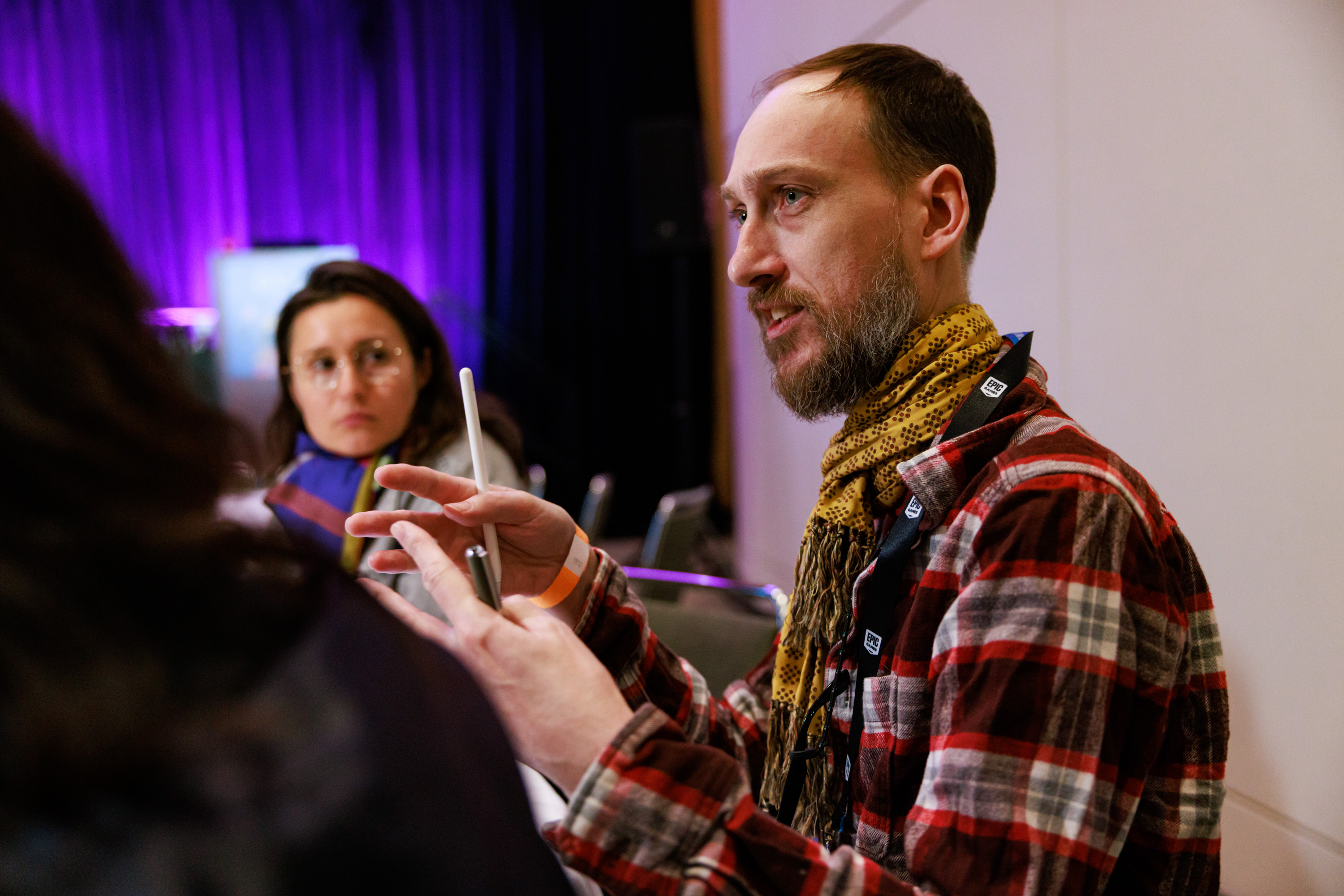 Adam Procter gestures with both hands discussing an idea during a round table session at GDC 2023, in his right hand he holds an Apple Pencil, in his left hand a uni-ball eye fine writing pen.